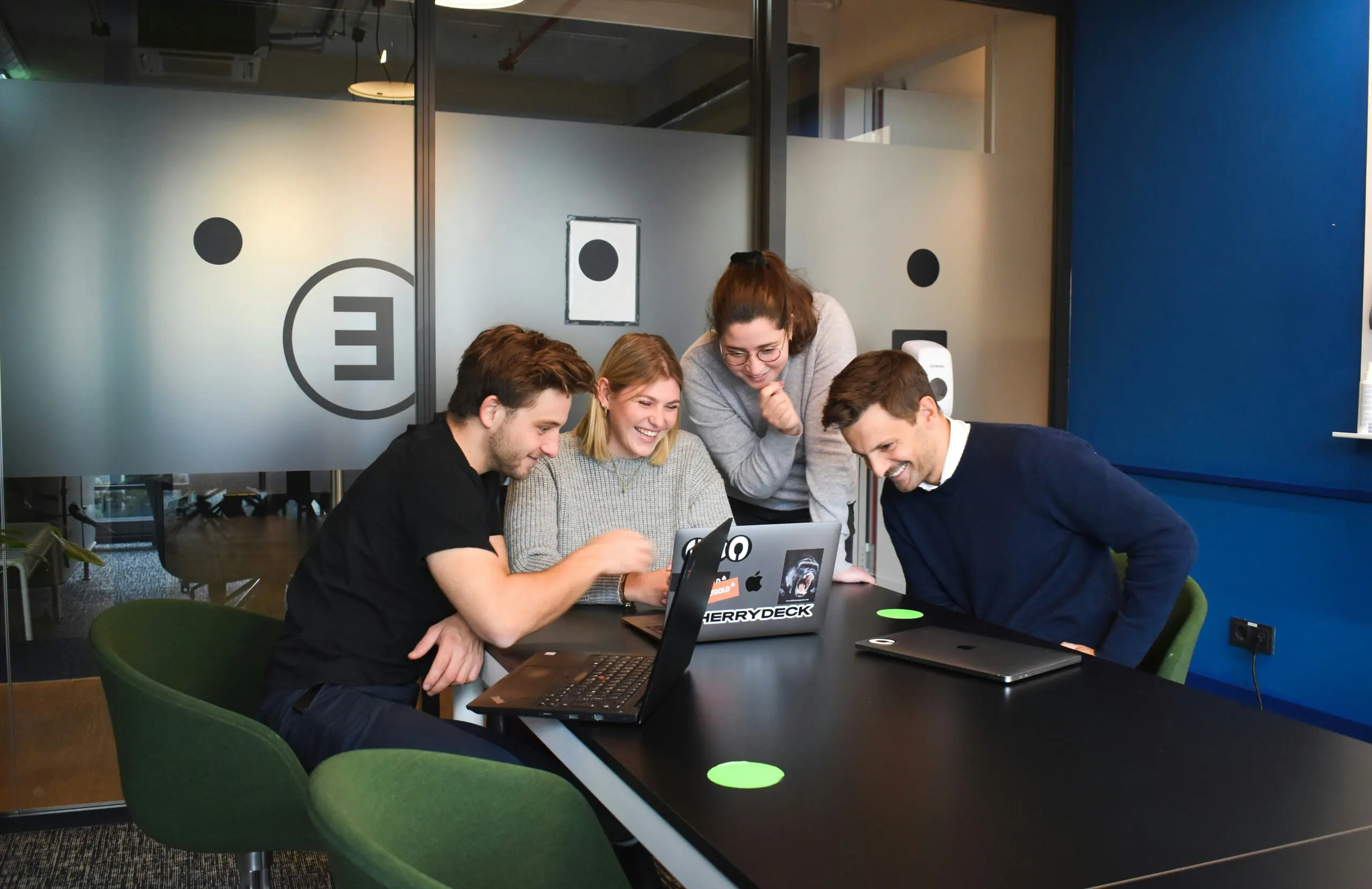 Four colleagues laughing together around a laptop at a meeting table