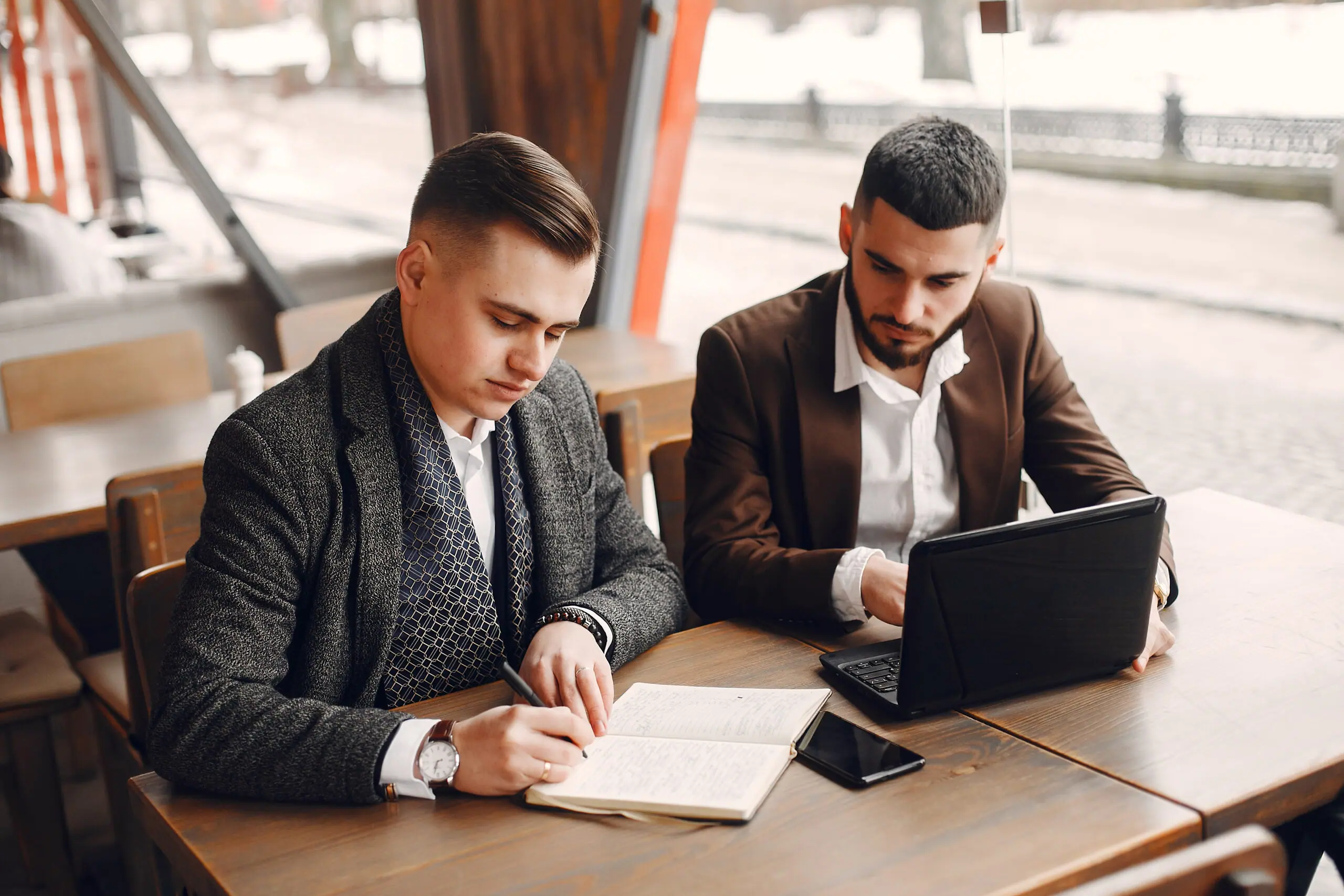 Two colleagues working at a café table with a laptop and notebook