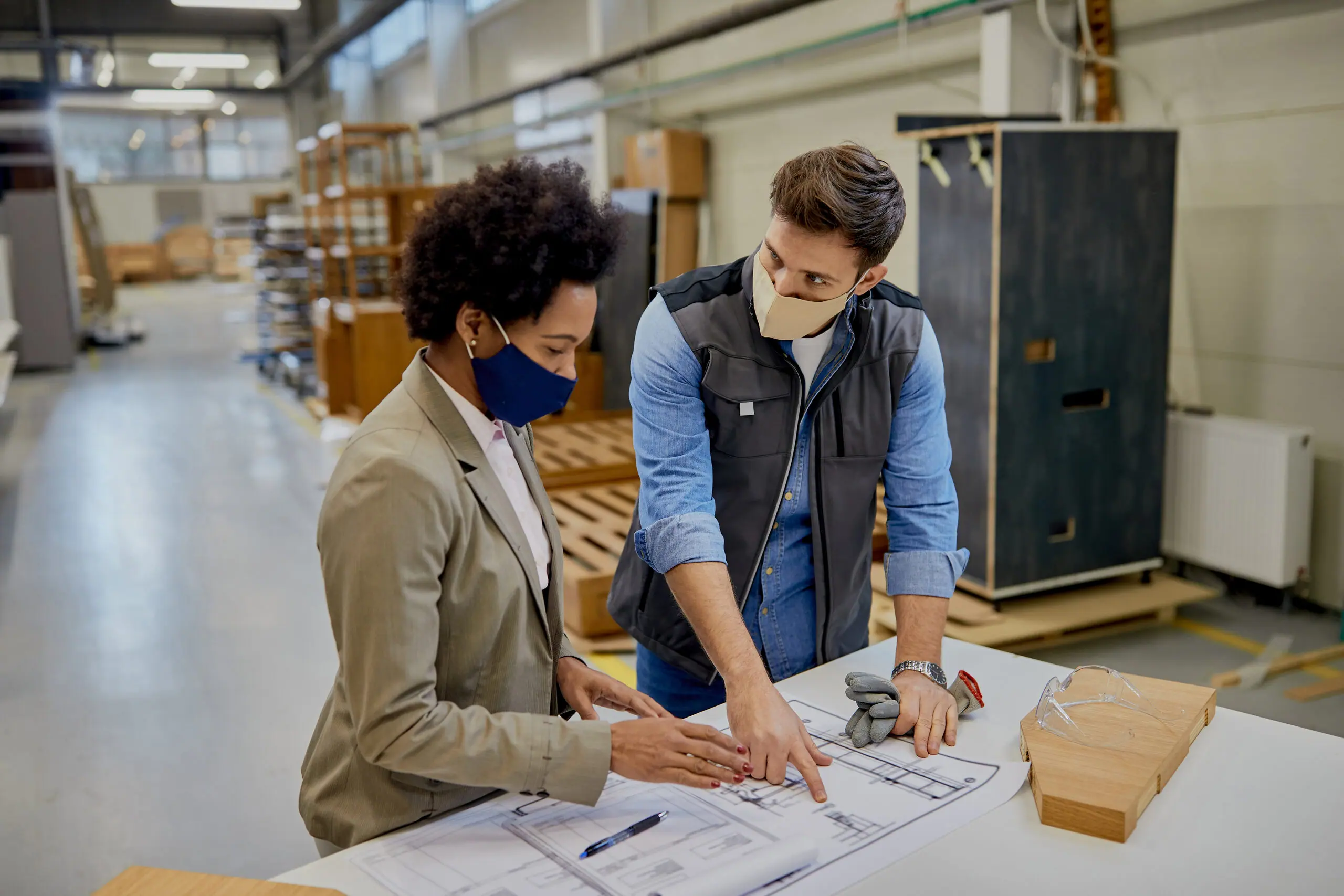 Two colleagues in face masks reviewing plans in a manufacturing workshop